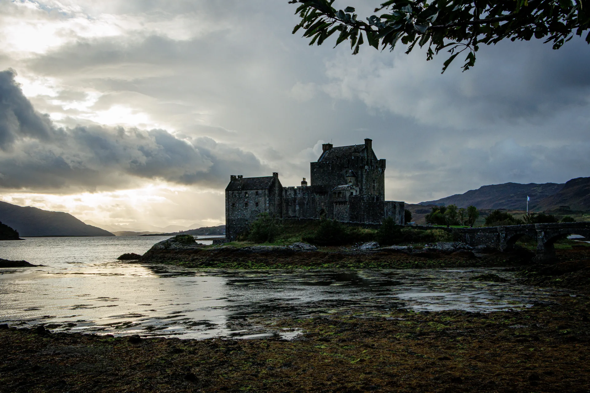 Eilean Donan Castle at low tide on Loch Duich, moody overcast sky with rays of light, Scottish saltire flying from the tower.