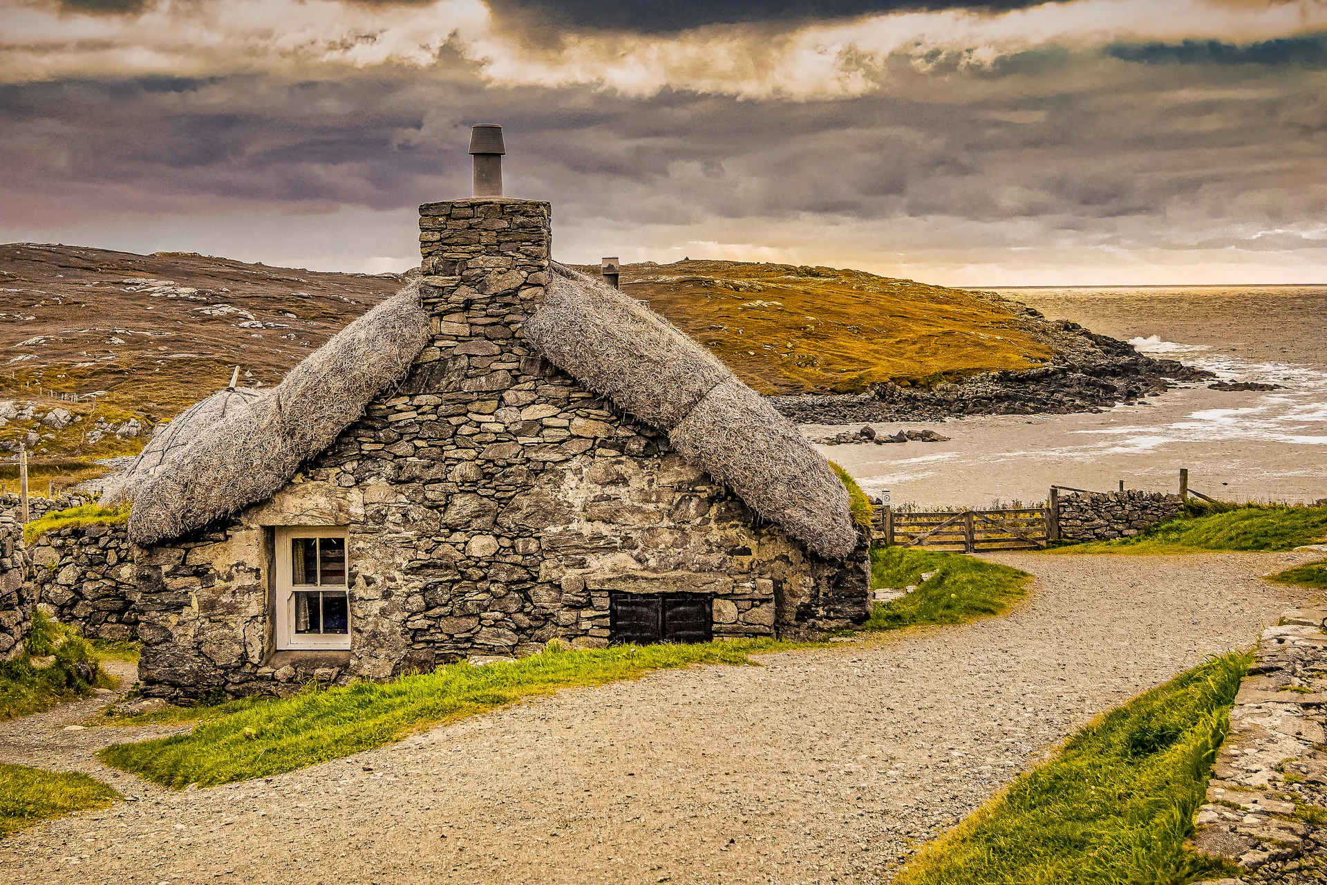 A restored thatched blackhouse cottage on the Isle of Harris, stone walls, dramatic storm sky, Atlantic sea in the background.