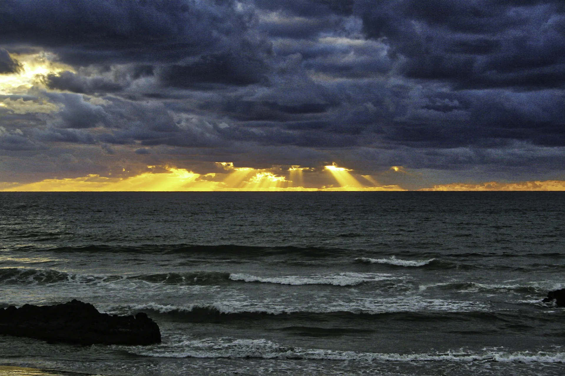 Crepuscular rays of light breaking through storm clouds over the Pacific Ocean from the Mexican coast, waves in the foreground.