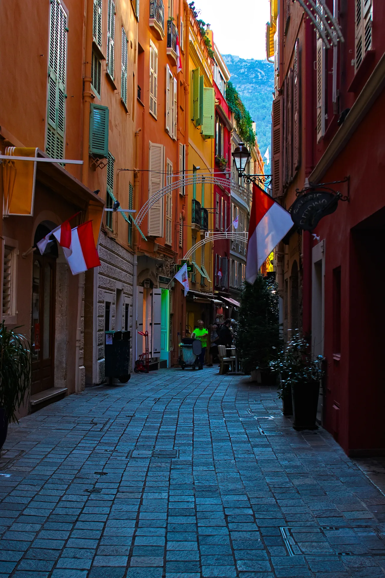 A narrow street in Monaco-Ville old town, warm ochre and terracotta buildings with Monegasque flags, cobblestones.