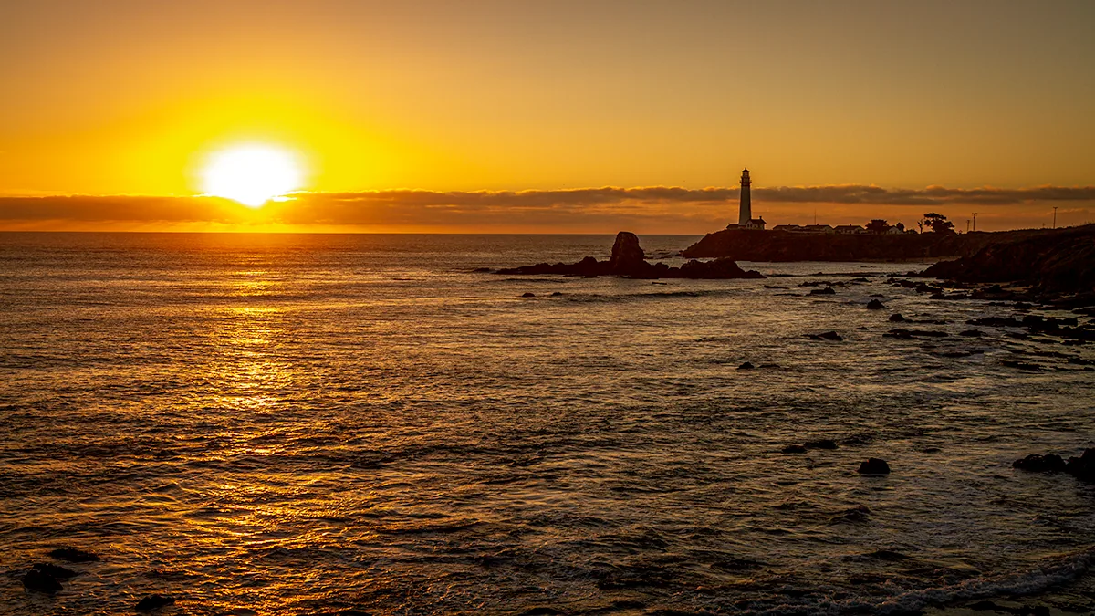 Pigeon Point Lighthouse silhouetted against a golden Pacific sunset, rocks in the foreground water.
