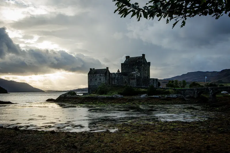 Eilean Donan Castle at low tide — moody overcast sky, Scottish flag, still water.