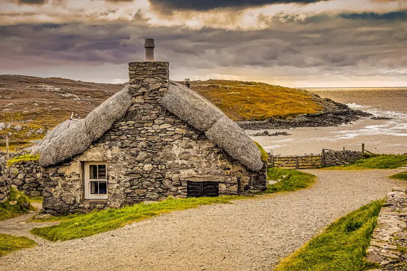 A restored thatched blackhouse cottage on the Isle of Harris, stone walls, dramatic storm sky, Atlantic sea in the background.