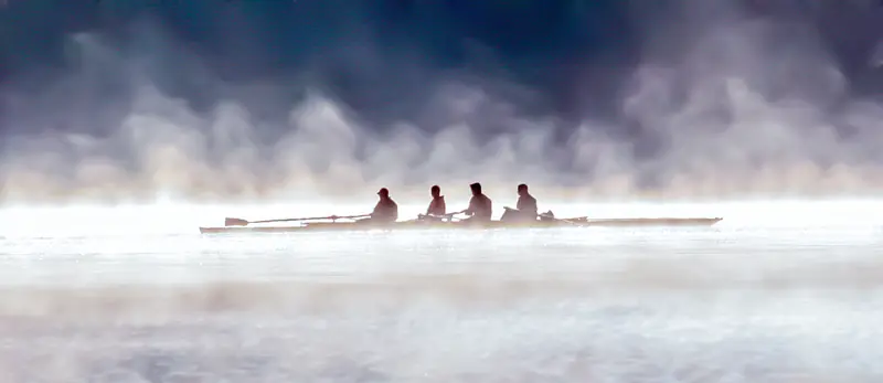 Four rowers in a shell emerging from morning mist on a river — ethereal silhouettes against white light.