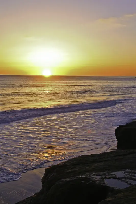 Pacific coast sunset from rocks — the sun's disc at the horizon, golden light on the waves, rock platform in the foreground.