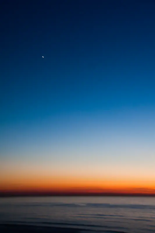 Minimalist twilight seascape — deep blue gradient sky, orange horizon band, sliver of moon.