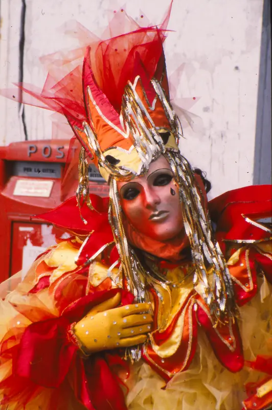 A Venetian carnival performer in a full red and gold costume and white mask, standing against a wall with a red letterbox.