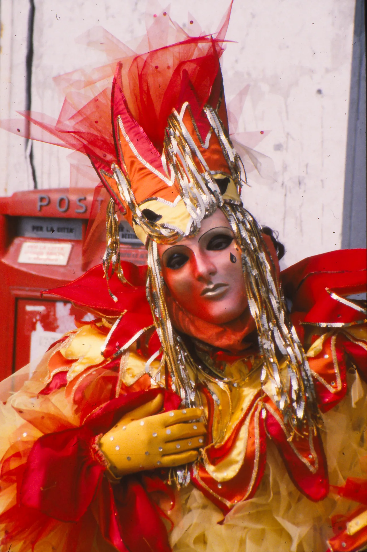 A Venetian carnival performer in a full red and gold costume and white mask, standing against a wall with a red letterbox.