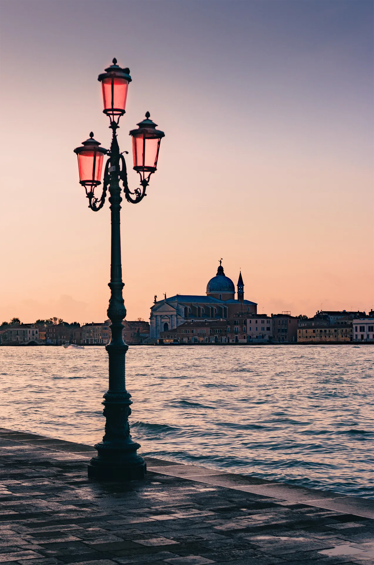 A Venetian street lamp with red glass on the Giudecca promenade at dusk, the Church of the Redentore across the lagoon.